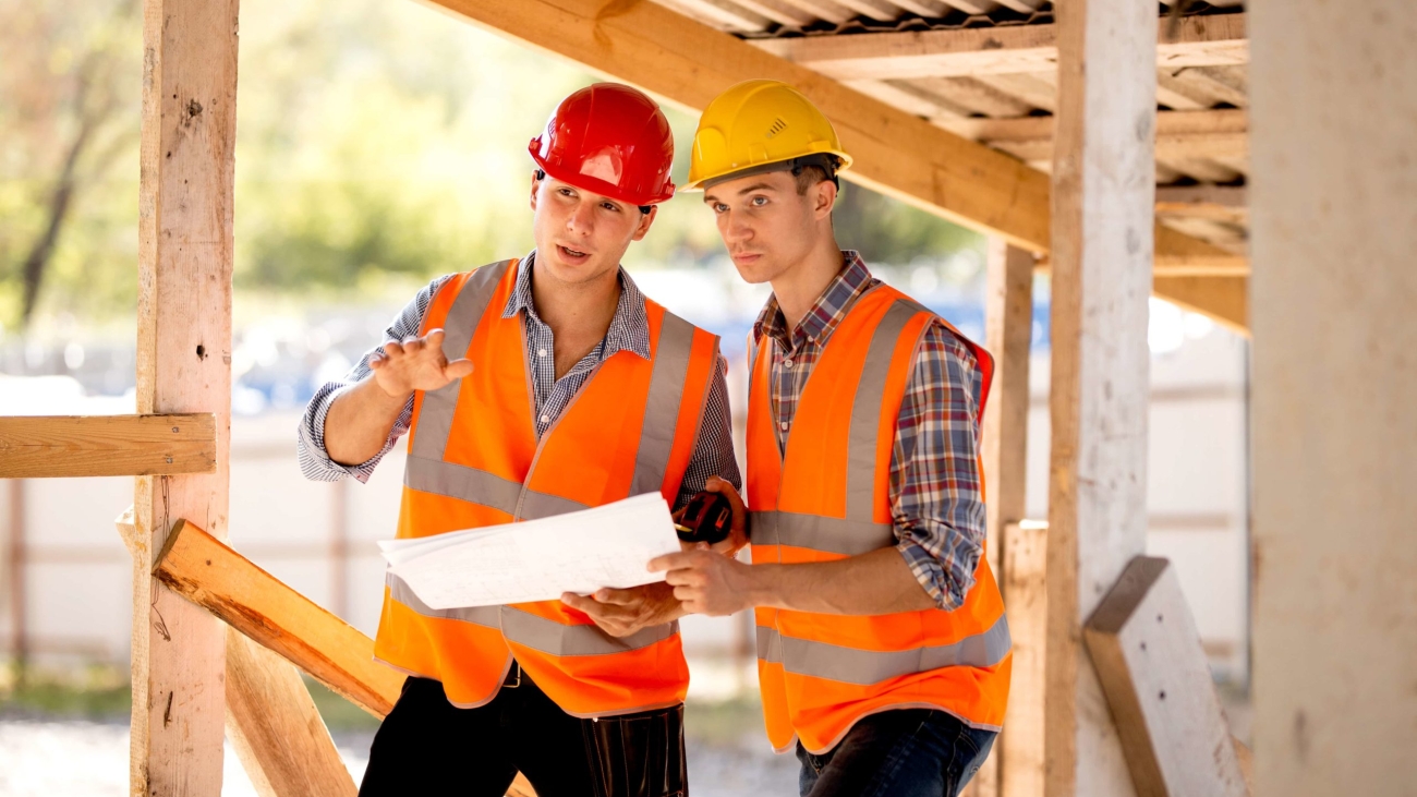 Two men dressed in shirts, orange work vests and helmets explore construction documentation on the building site near the wooden building constructions